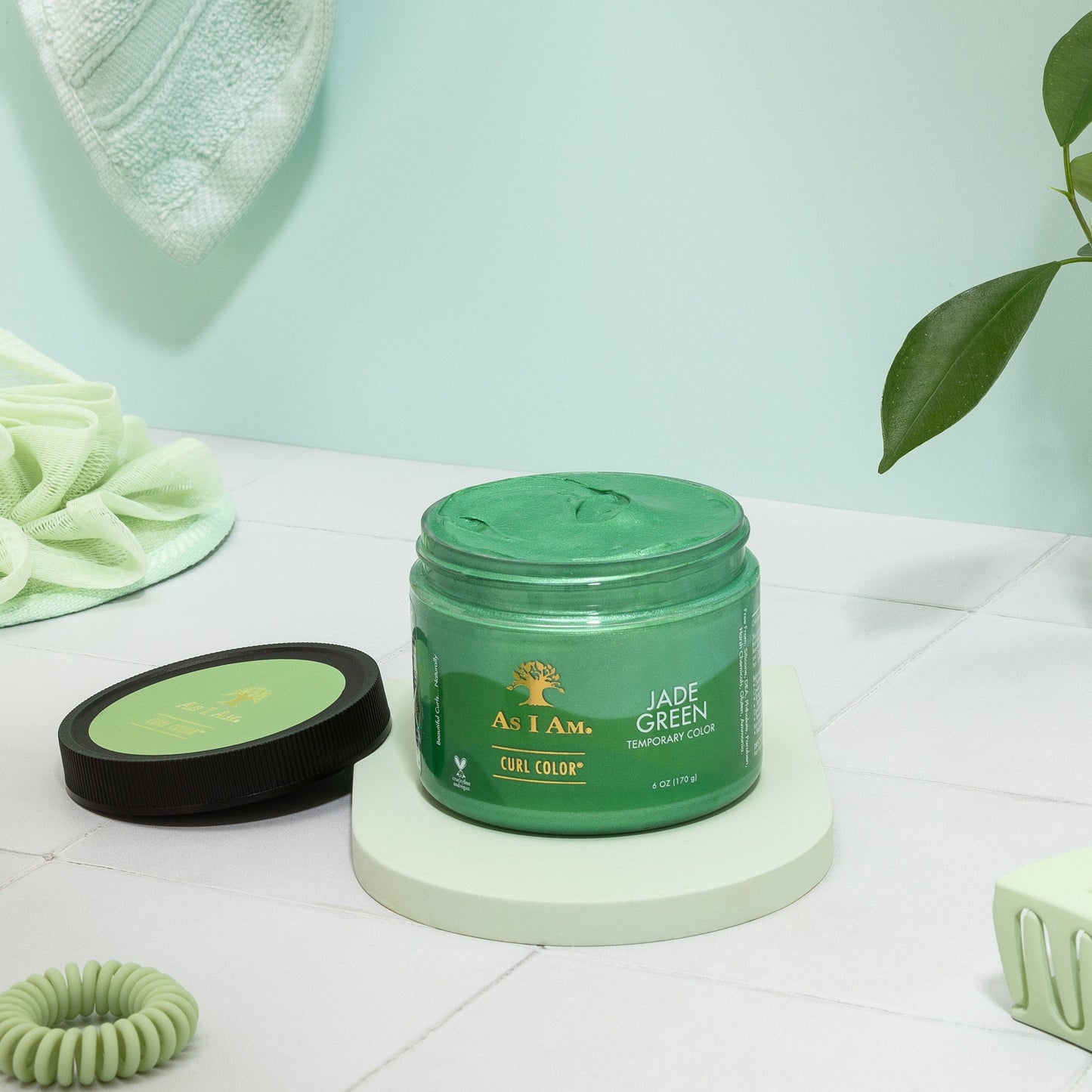 وعاء من كريم تجعيد الشعر بغطاء وملصق باللون الأخضر اليشم، معروض على سطح أبيض مع نبات في الخلفية.