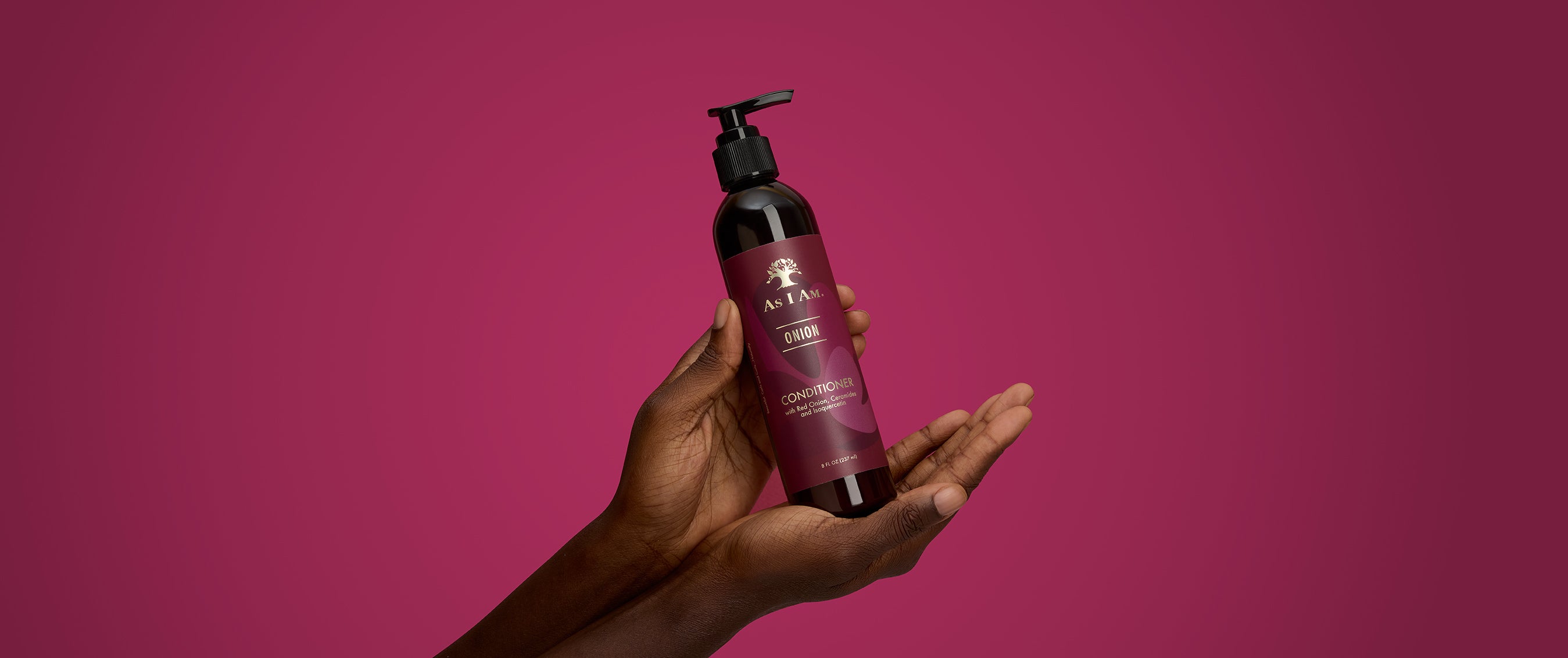 يد تحمل زجاجة من منتج العناية بالشعر "as i am" على خلفية وردية